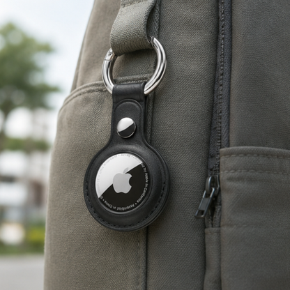 Apple AirTag secured in a black leather keychain case attached to the strap buckle of a dark backpack, shown outdoors in natural light