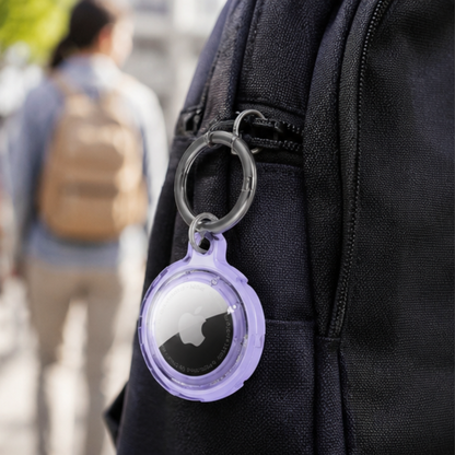 Apple AirTag in a clear transparent keychain case clipped onto the strap of a black backpack, shown in an outdoor lifestyle setting