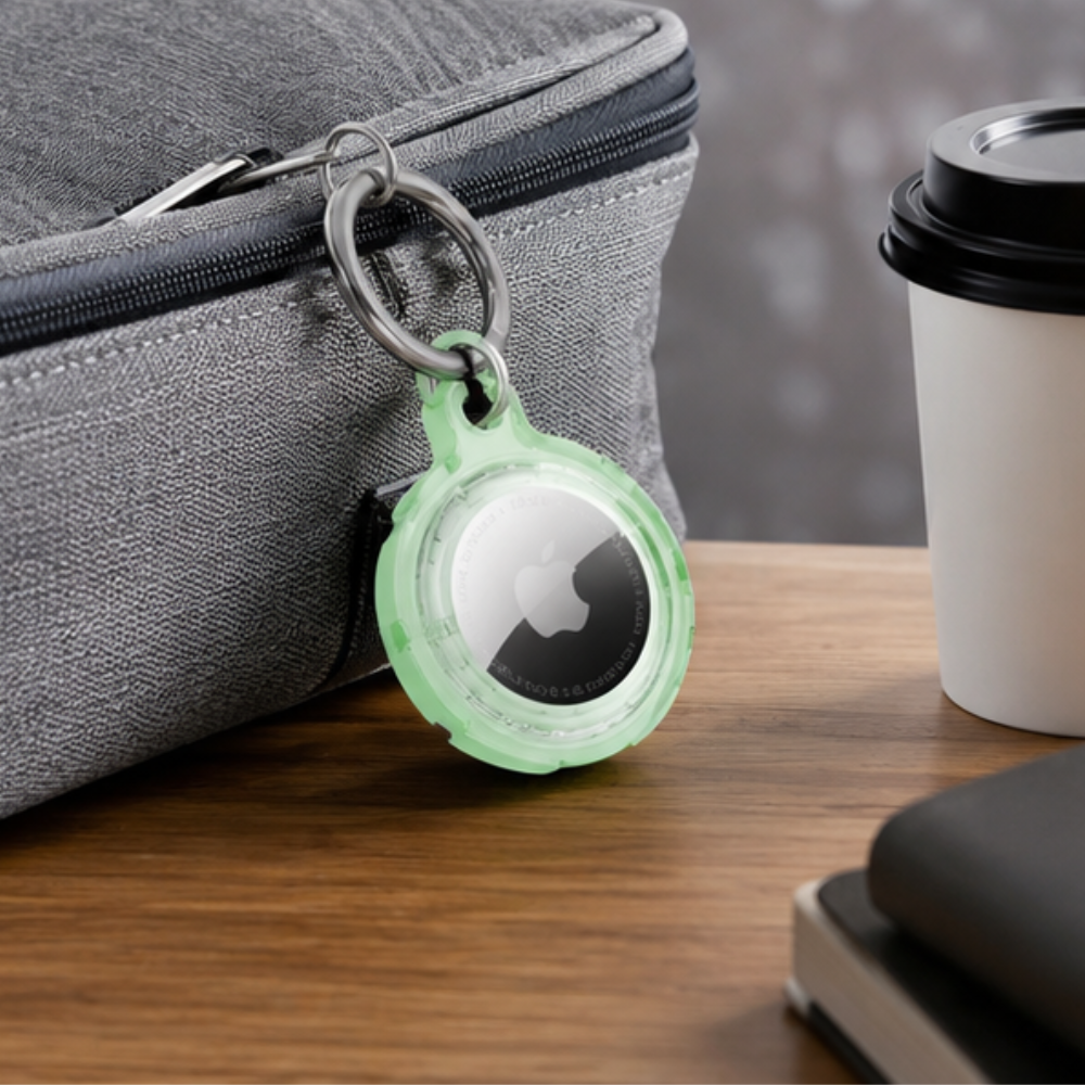 Apple AirTag in a mint green transparent protective case hanging from a gray bag zipper, displayed on a desk alongside a coffee cup