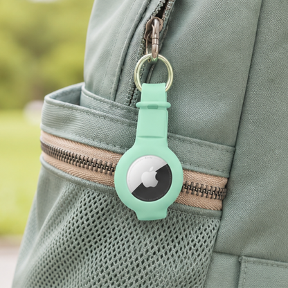 Apple AirTag in a mint green silicone keychain holder clipped onto the zipper of a sage green backpack, shown in an outdoor lifestyle setting