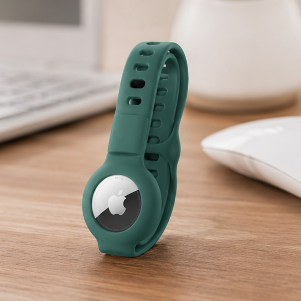 Apple AirTag secured in a dark green silicone wristband case displayed upright on a wooden desk surface, with a computer and keyboard visible in the background