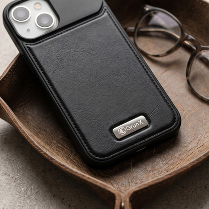 Black leather iPhone case with protective design displayed on wooden tray next to reading glasses and metal pen in premium workspace