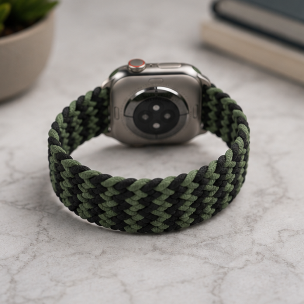 Apple Watch with dark green braided solo loop strap lying flat on a white marble surface with a plant in the background