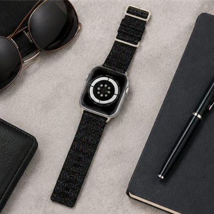 Apple Watch with a black woven nylon sport loop band displayed in a flat lay alongside sunglasses and a notebook on a gray textured surface