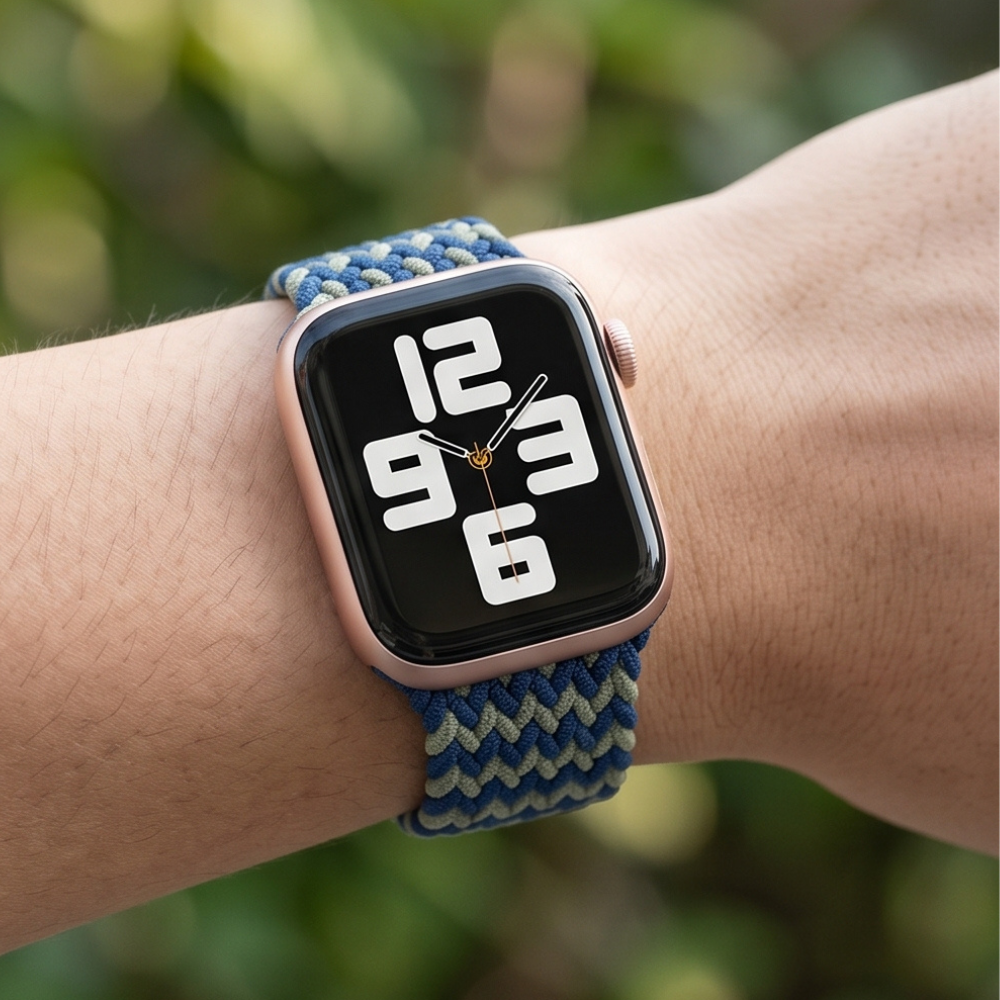 Close-up wrist shot of a person wearing an Apple Watch with a blue and green chevron braided solo loop band, shown against a softly blurred outdoor background