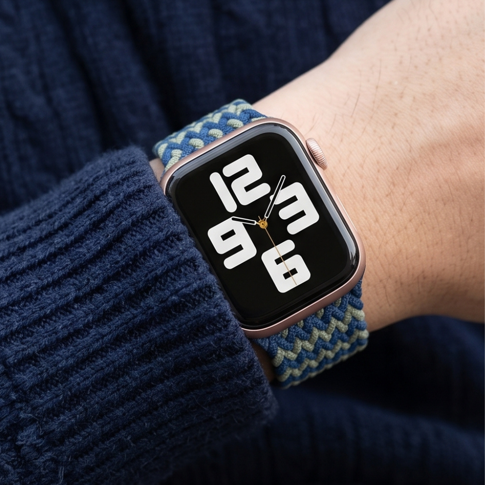Person wearing an Apple Watch with a navy blue and green zigzag patterned braided solo loop band on their wrist, styled with a dark knit sweater and coffee cup in the background