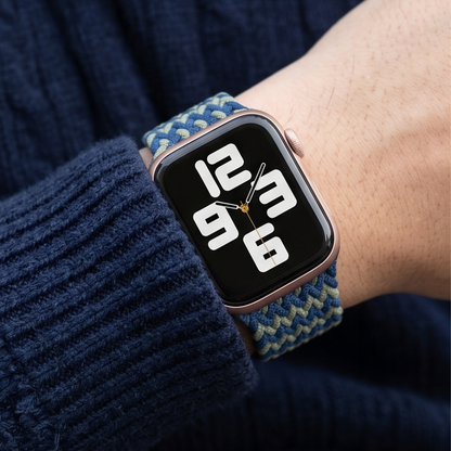Person wearing an Apple Watch with a navy blue and green zigzag patterned braided solo loop band on their wrist, styled with a dark knit sweater and coffee cup in the background