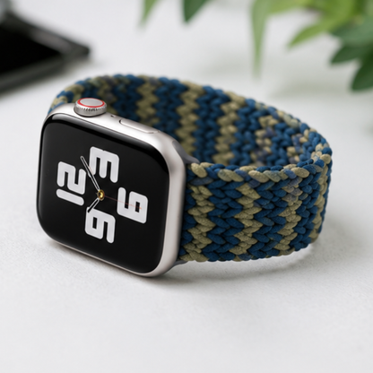 Apple Watch with a navy and green woven braided solo loop band placed flat on a dark gray surface next to a pair of white AirPods, in a tech lifestyle flat lay