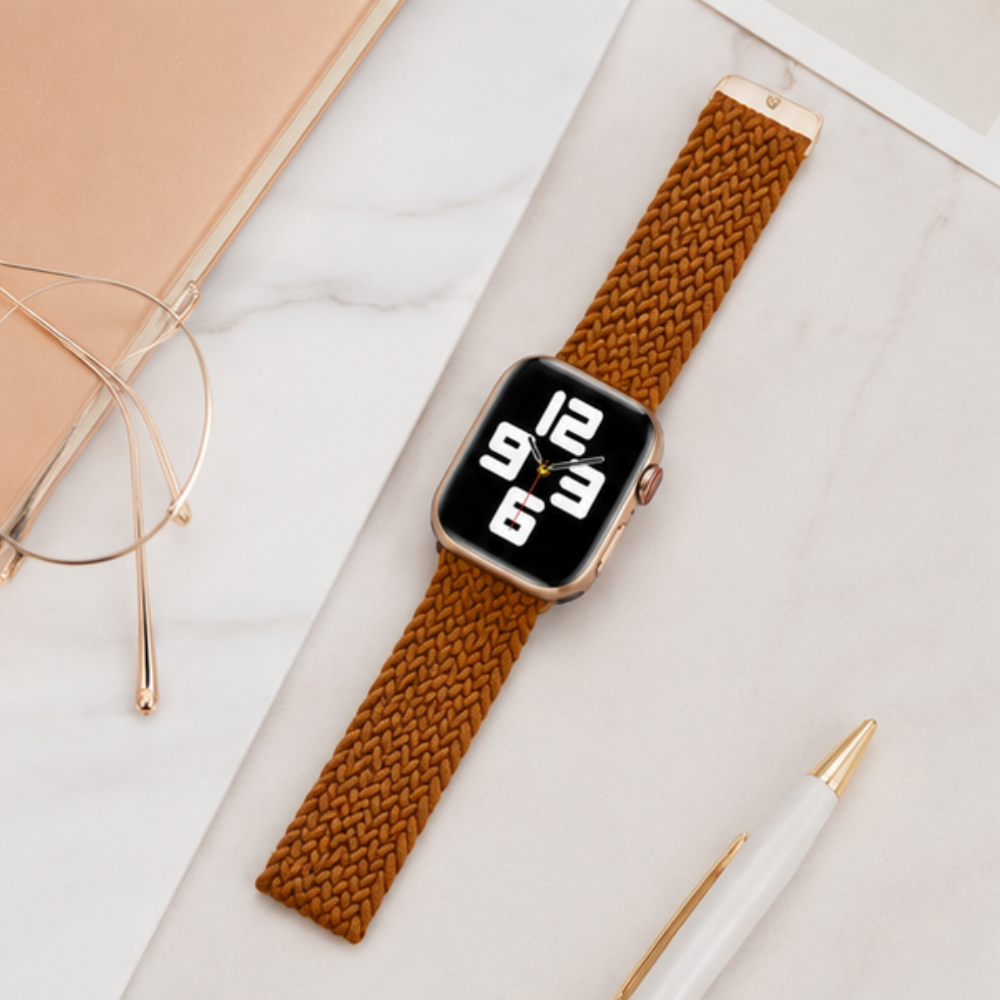 Apple Watch with a golden brown braided solo loop band laid flat on a white surface alongside glasses and a pen, styled in a workspace setting