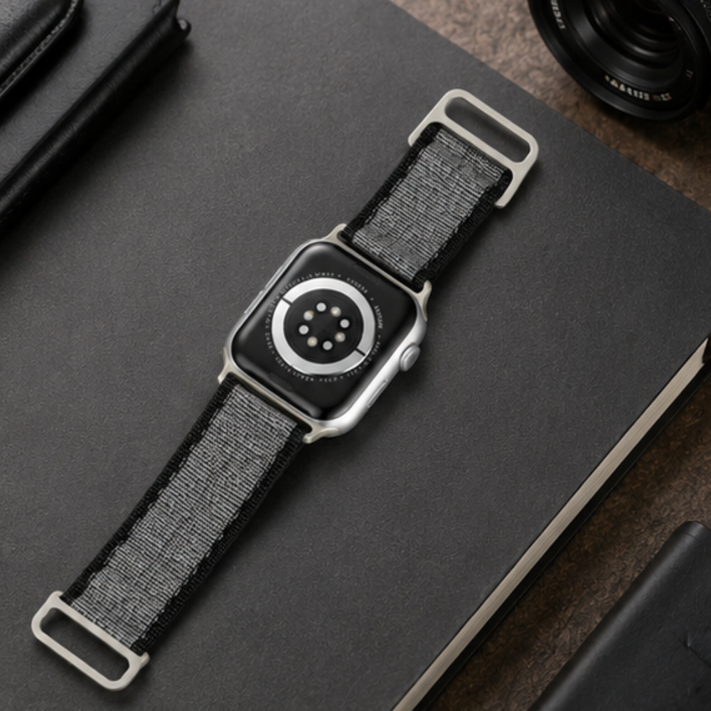 Apple Watch Ultra with charcoal grey woven nylon alpine loop band laid flat on a dark notebook beside a cable