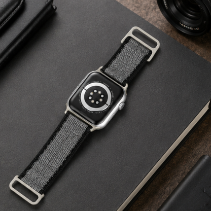 Apple Watch Ultra with charcoal grey woven nylon alpine loop band laid flat on a dark notebook beside a cable