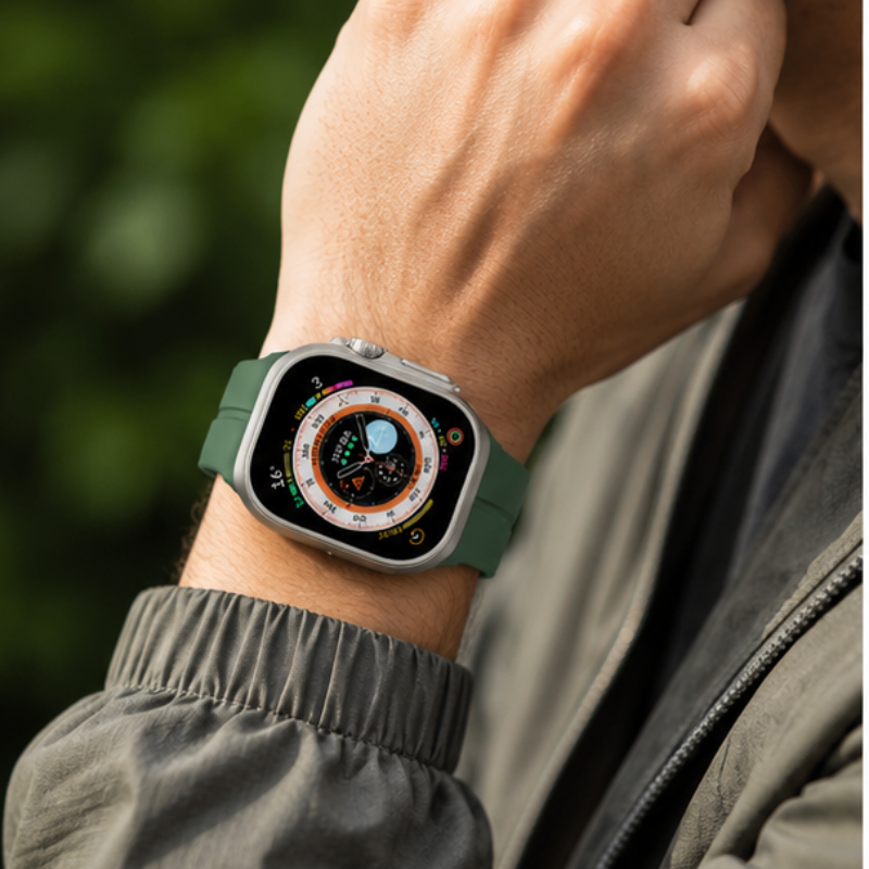 Person in a grey outdoor jacket wearing Apple Watch Ultra with forest green silicone sport band on their wrist against a blurred green nature background