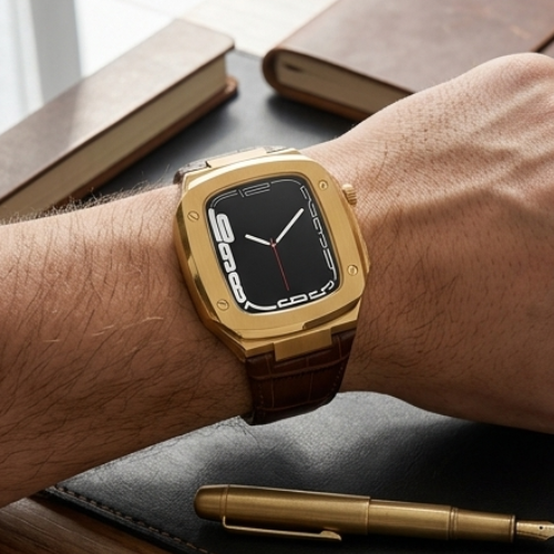 Apple Watch with gold case and black dial paired with brown crocodile-embossed leather band resting on a wooden surface beside a book