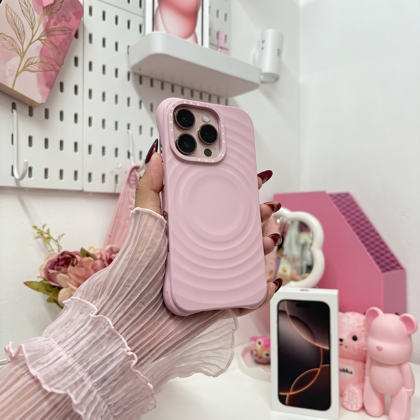 Pink wavy textured iPhone case with metal camera ring held in hand in aesthetic bedroom setting, surrounded by white pegboard organizer, pink flowers, and cute desk accessories