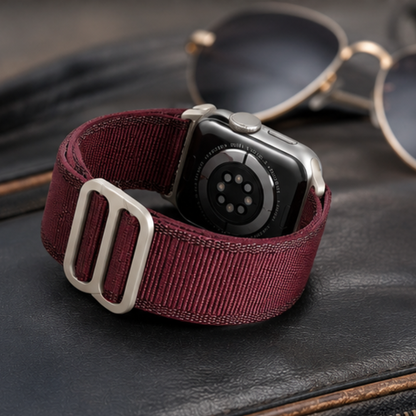Apple Watch Ultra with burgundy woven alpine loop band laid flat on a dark wooden surface beside sunglasses