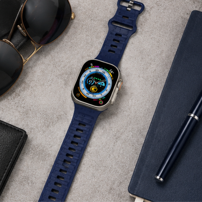 Apple Watch Ultra with a dark navy blue rubber band placed on a flat lay arrangement with sunglasses, a pen, and a notebook on a dark surface