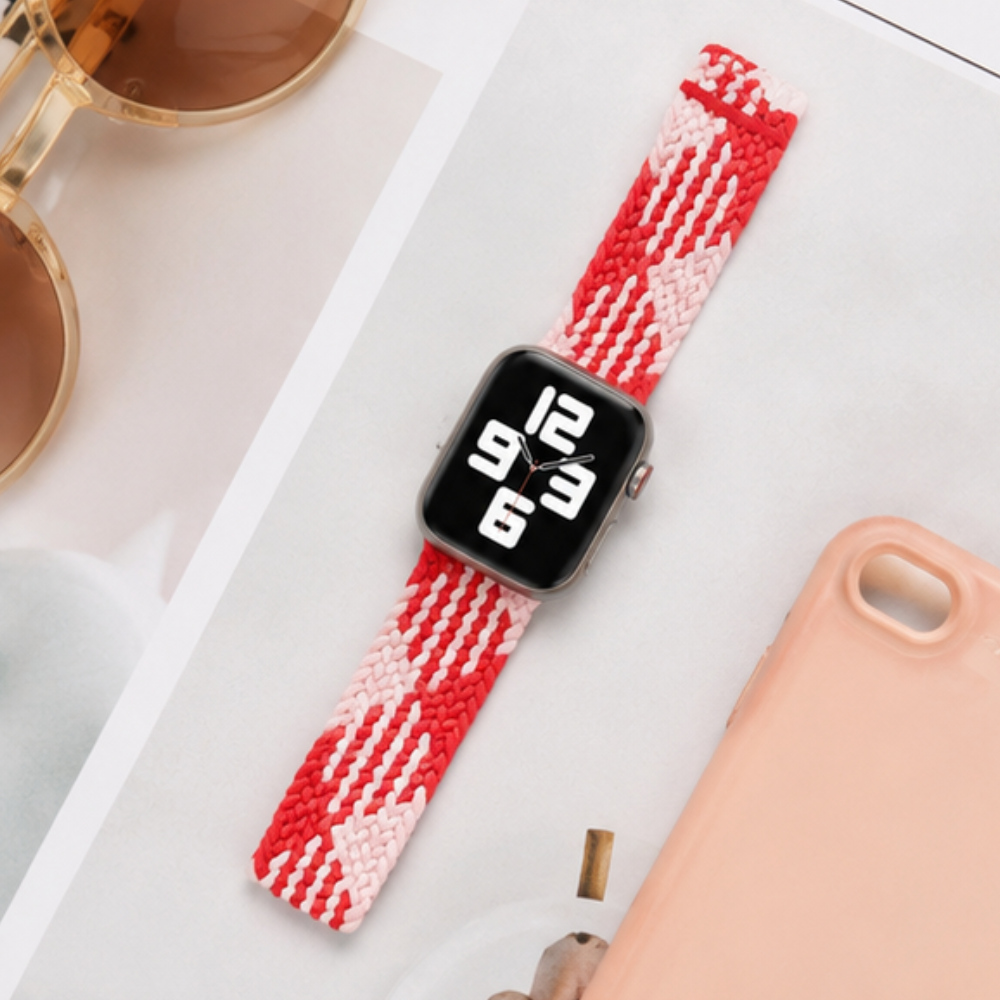 Apple Watch with a red and white zigzag patterned braided solo loop band laid flat on a white surface next to a pink phone case