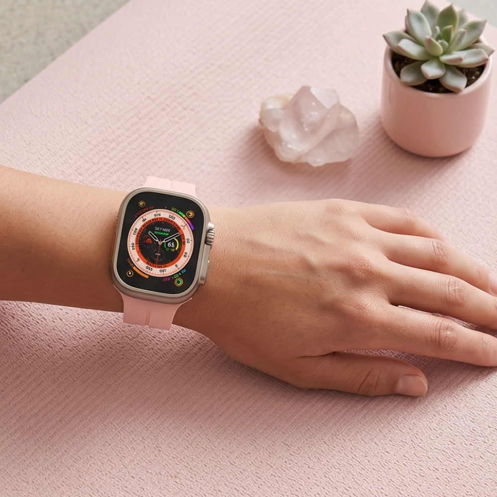 Pink edition smartwatch with orange accent ring worn on wrist on pink desk surface, styled with succulent plant, white candle, and minimalist workspace