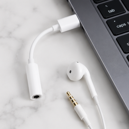 White USB-C to 3.5mm audio adapter connected to a laptop with earbuds on marble surface