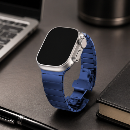 Apple Watch Ultra with silver case and blue stainless steel link band placed on a dark notebook beside a pen and laptop