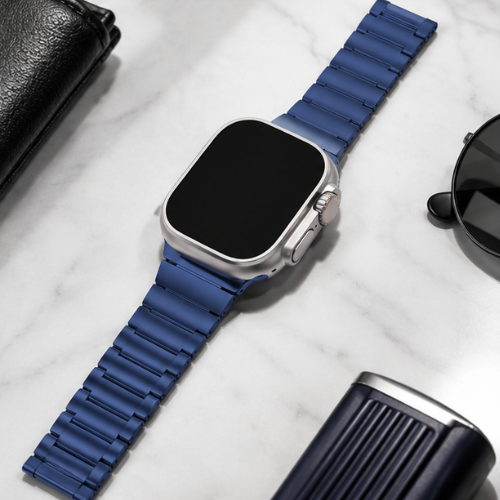 Apple Watch Ultra with silver case and blue stainless steel link band laid flat on a white marble surface beside sunglasses and a leather wallet
